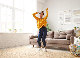 Young playful african american woman in headphones jumping and dancing in living room at home