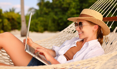 Working at sunset. Side view of young happy female using laptop while lying in the hammock on the beach