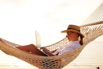 Working at sunset. Side view of young happy female using laptop while lying in the hammock on the beach