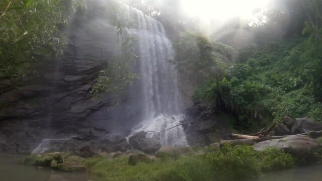 A waterfall in the jungle with a heavy mist blowing in the wind and the sun shining in the sky.