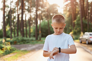 Little blonde boy using fitness bend touching button and touchscreen while posing outdoor in park, touching button and setting smart watch before running on road.