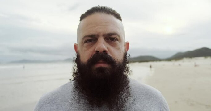 Serious latin man with big beard on a beach looking at horizon. 4K.