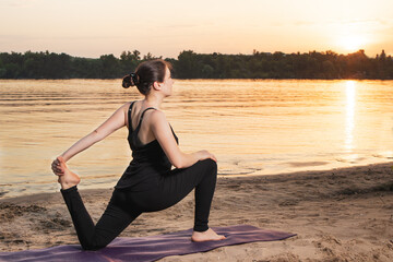 A woman in sportswear practices yoga at sunset near the river on the beach in nature. Stretching the front of the thigh of the quadriceps, place for text