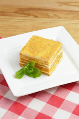One piece of honey cake on a plate with mint leaf on top. Served on a white plate over red plaid background.