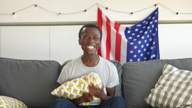Front shot of a young afro-american guy sitting on the sofa at home looking at the camera and exults for success after reading the good news.