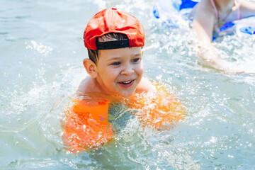 Happy kid in a red cap and inflatable life jackets on his arms swims in the water. Children splash in the water outdoors in the summer. Children's active rest