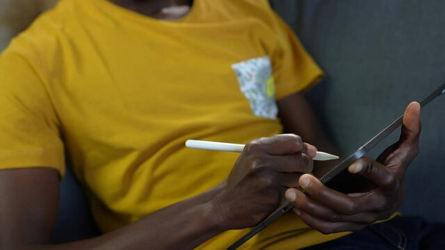 Closeup on black afro american young man's hands using a digital tablet and a smartpen on the screen to work or study on the sofa at home.
