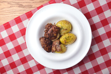 Grilled meat with baked potatoes. Served on a white square plate over red plaid tablecloth.