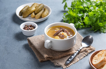 Pickle (Russian soup). Pearl soup with pickles and potatoes in a white plate on a blue background with bread and fresh  herbs. Copy space