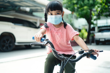 Asian school kid girl in medical mask riding a bike or bicycle on a holiday. Happy child biking during coronavirus covid-19 epidemic.