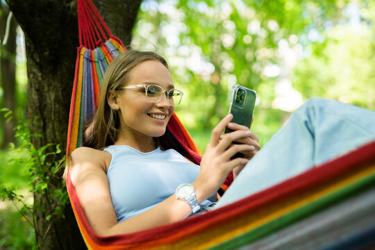 Young Woman Chilling In Hammock And Holding Phone Girl With Phone Chatting With Friends Or Making Order Online, While Relaxing In Hammock.