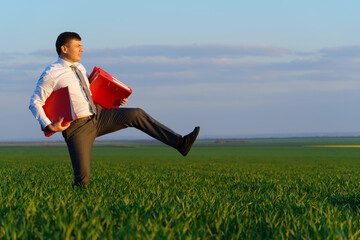 businessman walks through a green grass field and holds an office red folder with documents - business concept