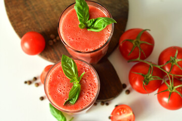 Two glasses of fresh organic tomato juice decorated with raw tomatoes and green leaves of basil on light background.Healthy diet nutrition concept.