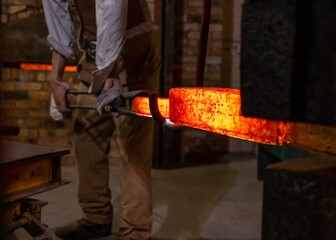 Foundry worker old blacksmith out of focus metal in focus protective clothing forming steel from orange molten metal. Forging glowing red hot iron in industrial setting with pressure press machine.