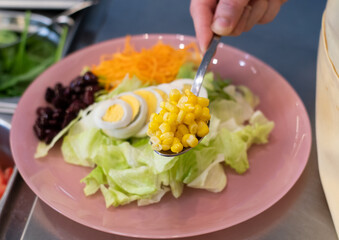cropped view of woman hand adding corn in pink plate with salad in restaurant. Serving healthy vegetarian salad