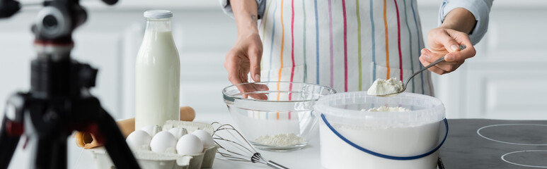 partial view of woman in apron holding spoon with flour near blurred digital camera, banner.