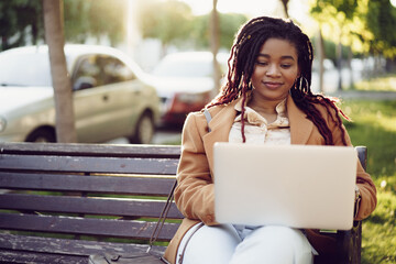 African american woman freelancer sitting on a bench in a street and using laptop