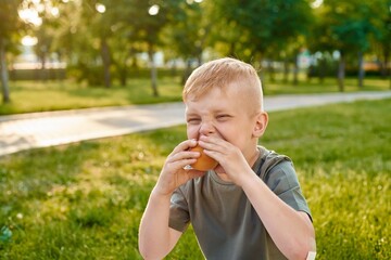 A small five-year-old red-haired boy eats a juicy orange peach in the park, close-up