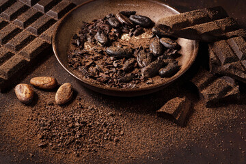 Chocolate and cacao concept. Cocoa powder in bowl near cocoa beans and broken chocolate on black background