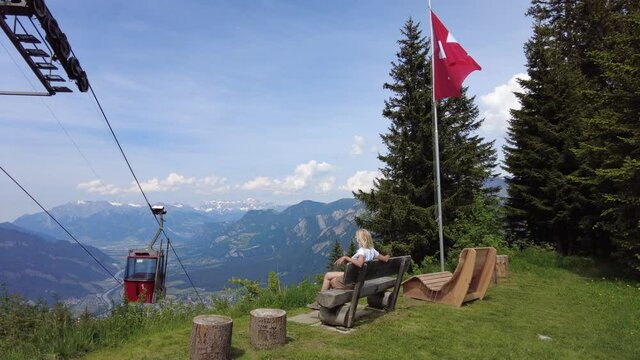 Woman relaxing on top of Brambruesch location in Switzerland. Swiss cable car of Chur or Coira with Swiss flag. Chur skyline in Grisons canton. Red cable car cabin from Chur to Kanzeli and Brambruesch