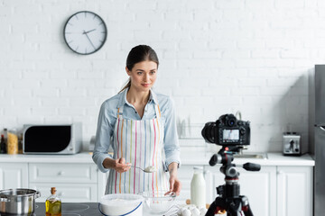 young culinary blogger holding spoon with flour in front of blurred digital camera.