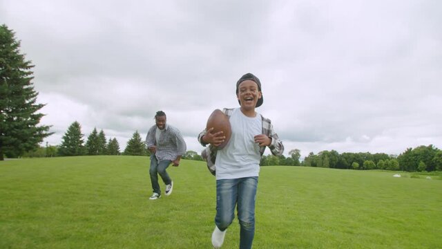 Excited happy preadolescent african american boy carrying ball,, running through green field to score touchdown, chased by carefree father while joyful family playing american football outdoors.