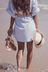 An unrecognizable black girl strolling along the beach holding her hat and flip-flops while she is...
