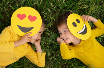 Two funny little girls are lying on the lawn covering part of their faces with emoticons and...
