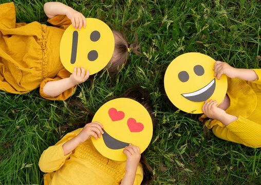 Three Children Lying On The Grass Are Holding Cardboard Emoticons With Different Emotions In Their Hands: A Sad, Smiling Happy Smile, A Loving Smiley With Hearts Instead Of Eyes.  World Emoji Day