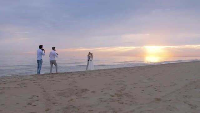 wide shot of the beach with the sun setting over the sea, of two photographers shooting a newlywed couple during their wedding