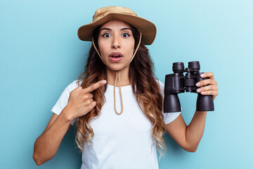 Young mexican woman holding binoculars isolated on blue background pointing to the side