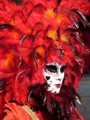 Parure vénitienne de plumes rouges autour d'un masque blanc aux lèvre écarlates. La pupille transparente en profondeur met en valeur le diadème du personnage travesti. Emblème du Carnaval de Venise