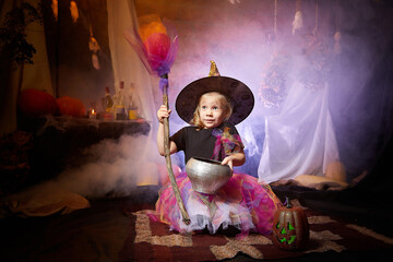 Little cute blonde girl looking as witch in special dress and hat in room decorated for Halloween. Witchcraft and wizardry in carnival. Halloween style photo shoot.