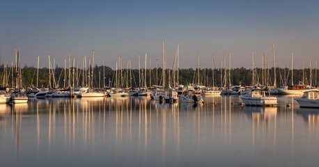 A marina of sailing boats at sunrise with numerous reflections of their masts all on the Beaulieu River at Buckler's Hard, Hampshire UK - obrazy, fototapety, plakaty