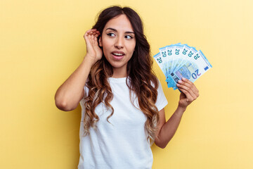 Young mixed race woman holding bills isolated on yellow background trying to listening a gossip.