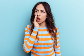 Young mexican woman isolated on blue background is saying a secret hot braking news and looking aside