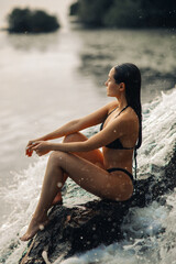Fototapeta premium Woman sits under the waterfall and looks at the river.