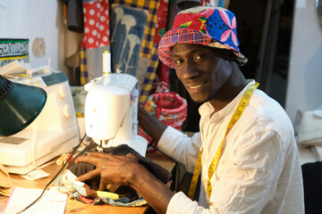 African tailor sewing a hat with sewing machine at sewing workshop smiling and looking at camera. Black man working with african fabrics.