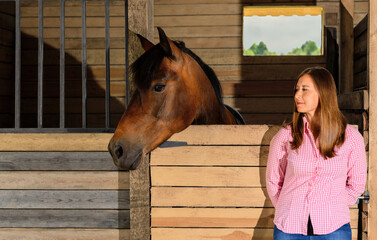 Caucasian woman and her bay horse are standing in a stable.