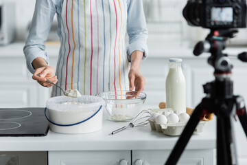 cropped view of woman in apron adding flour into bowl near blurred digital camera.