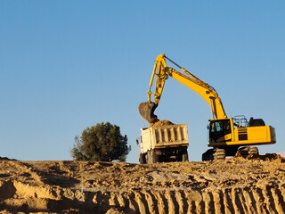 Excavator machine filling truck in construction site on sky background.