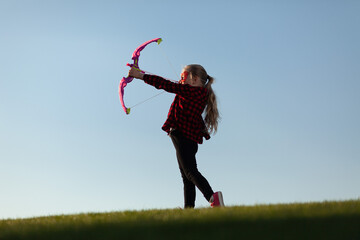 Little girl shoots a bow on the background of the horizon. Imaginary archer game, superhero in...