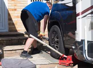 A mechanic uses a metal pole on a wrench to remove the wheel nuts from a motorhome wheel on a private drive.Trolley jack visible in foreground.Man wears shorts