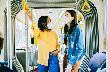 Two cheerful pretty young multiethnic women in protective masks are standing in a bus and looking...
