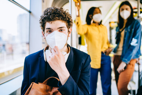 Portrait Of Young Tired Handsome Caucasian Man With Medical Mask FFP2 KN95 Looking At Camera Sitting In Bus With Other Passengers On Blurred Background. Train Passenger Traveling Safely In Seat.