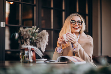 Elderly business woman in a coat sitting outside cafe and reading magazine