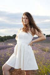 Fototapeta na wymiar Portrait of a young woman in the lavender fields of Brihuega, in Spain. She wears a white dress and her hair is loose and curly. She is with her arms around her waist. Lifestyle. Beauty. fashion.