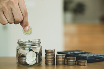 Businessman hand putting money coin stack in the jar placed on a wooden table saving for the future growing business, education and retirement .
