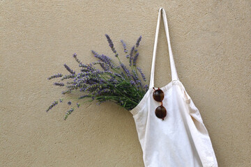 Canvas tote bag filled with fresh lavender flowers and retro sunglasses, hanging on a concrete wall.