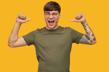 Fototapeta premium Young handsome male blogger, smiling broadly, points with both thumbs at himself, wink and feels happy. Wears green t-shirt and round glasses. Isolated over yellow background.